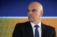 Retrato do ministro do Supremo Tribunal Federal (STF) Alexandre de Moraes, um homem branco e careca, de terno preto, camisa branca e gravata azul escura com padrões discretos está em frente a uma tela digital azul e laranja.