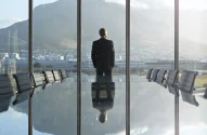 Um homem de costas em um terno preto, olhando pela janela de uma moderna sala de reuniões, com vista para uma cidade e montanhas ao fundo, em um momento contemplativo.