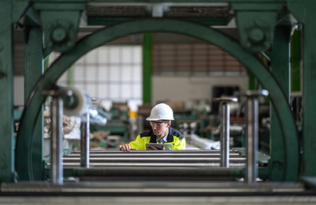 Trabalhador com capacete e colete refletivo em ambiente industrial, visto através de máquina.