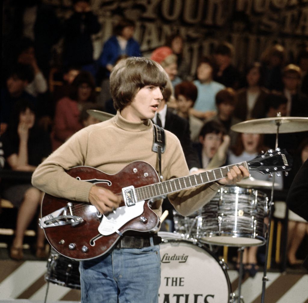 George Harrison dos Beatles tocando guitarra e cantando em palco.
