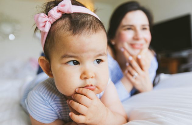 Bebê de laço rosa de bruços na cama, com a mãe desfocada sorrindo ao fundo.