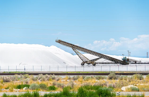 Em um cenário ensolarado, uma esteira transportadora se destaca no centro de uma paisagem industrial com montes de materiais brancos, cercada por trilhos de trem e vegetação rasteira.