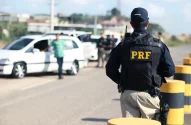 Na imagem, um policial da PRF de costas, com uniforme preto e inscrições amarelas, observa uma operação de fiscalização em uma estrada com barreiras, veículos parados e outros policiais. Ao fundo, construções e uma área verde sugerem tráfego na região.