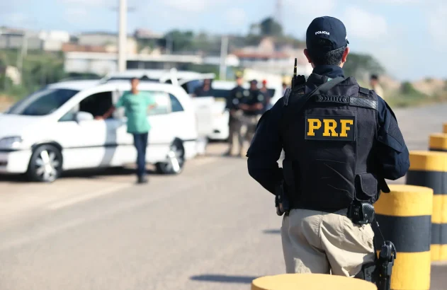 Na imagem, um policial da PRF de costas, com uniforme preto e inscrições amarelas, observa uma operação de fiscalização em uma estrada com barreiras, veículos parados e outros policiais. Ao fundo, construções e uma área verde sugerem tráfego na região.