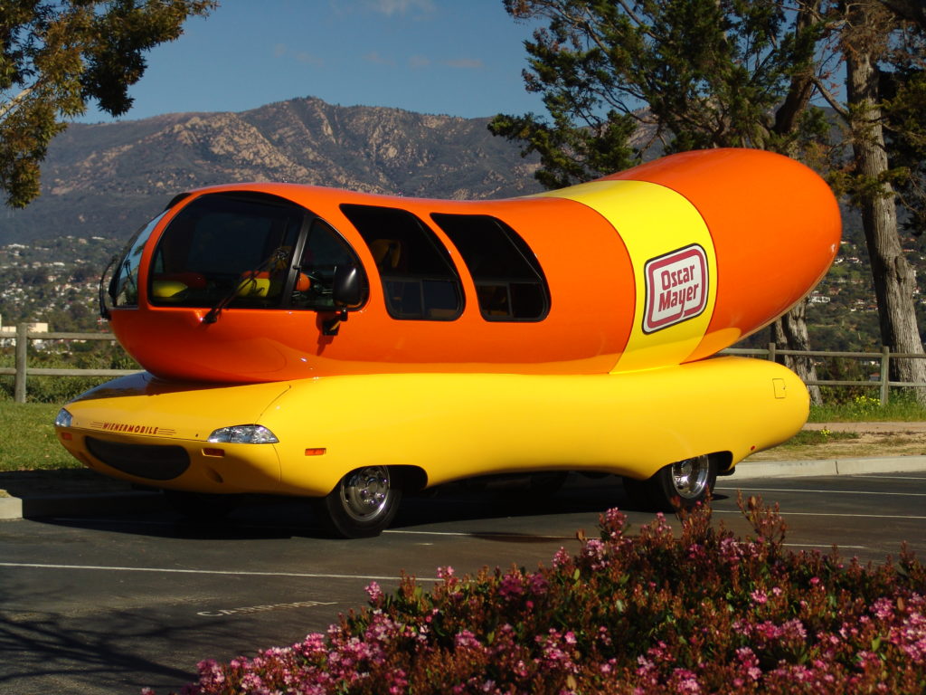 Oscar Mayer Wienermobile, um veículo gigante em forma de cachorro-quente laranja e amarelo, estacionado em área com montanhas.