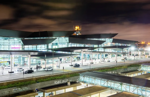 Entrada do Aeroporto de Guarulhos, com estruturas de vidro, torre de controle iluminada, telhado dinâmico, via movimentada e iluminação intensa.