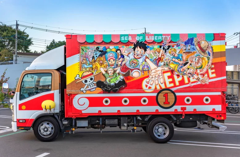 Um caminhão com carroceria colorida retratando personagens de One Piece em poses dinâmicas, com fundo vermelho, detalhes dourados e verdes, e a inscrição "SEA OF LIBERTY" no toldo superior, estacionado em um dia claro.