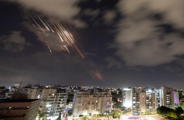 Céu noturno sobre cidade. Múltiplos rastros de luz brilhante de projéteis ou defesa aérea cortam as nuvens.