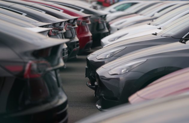 Fileiras paralelas de carros modernos estacionados em pátio. Veículos pretos, brancos, cinzas e vermelhos, vistos de frente e trás.