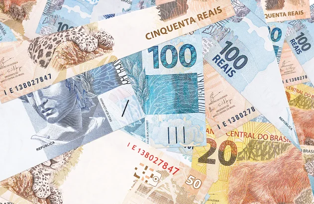 Um monte de cédulas de real espalhadas, destacando as notas de 50, 100 e 20 reais com imagens de uma onça-pintada, um peixe e um mico-leão-dourado, respectivamente.