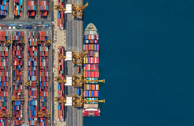 Vista aérea de um porto com contêineres coloridos organizados à esquerda, guindastes amarelos no centro-esquerdo, um navio cargueiro atracado à direita e o mar azul profundo ao fundo.