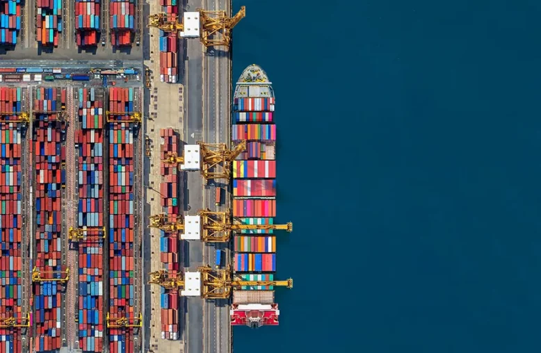 Vista aérea de um porto com contêineres coloridos organizados à esquerda, guindastes amarelos no centro-esquerdo, um navio cargueiro atracado à direita e o mar azul profundo ao fundo.