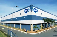 Fachada de um grande edifício industrial nas cores branco e azul com logos azuis da "EMS", palmeiras ao longo da calçada e céu azul ao fundo.