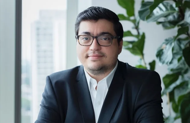 O economista Felipe Salto, um homem de cabelo curto e escuro, usando terno preto e óculos, está em um escritório com janela grande e uma planta verde ao fundo.