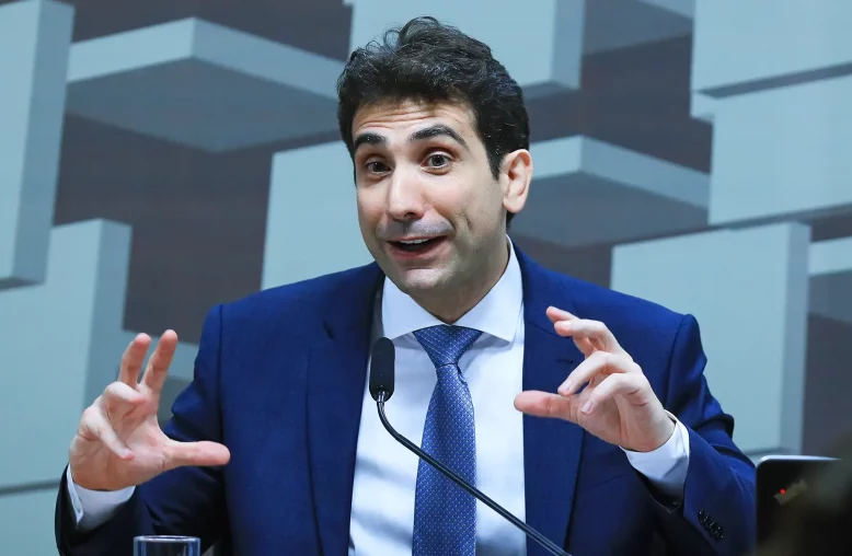 Retrato do economista Gabriel Galípolo, um homem de cabelo escuro, vestindo um terno azul escuro, camisa branca e gravata azul clara, gesticulando enquanto fala ao microfone com um fundo de figuras geométricas cinza.