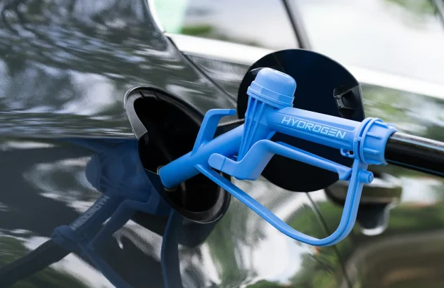 Bico de abastecimento de hidrogênio verde conectado a um veículo de cor escura, refletindo árvores e luz solar.