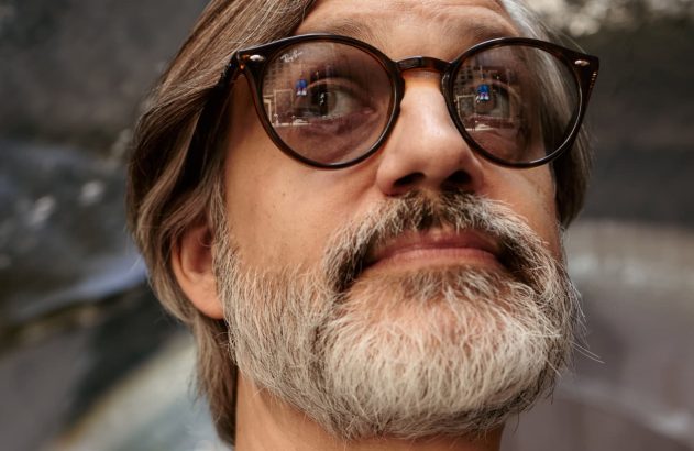 Close-up de homem barbudo de óculos, com olhos arregalados, olhando para cima e para a direita.