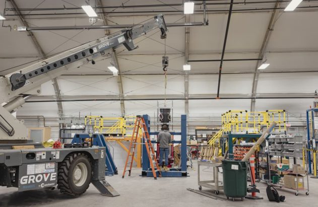 Guindaste Grove levanta peça; operário trabalha em equipamento em grande hangar industrial.