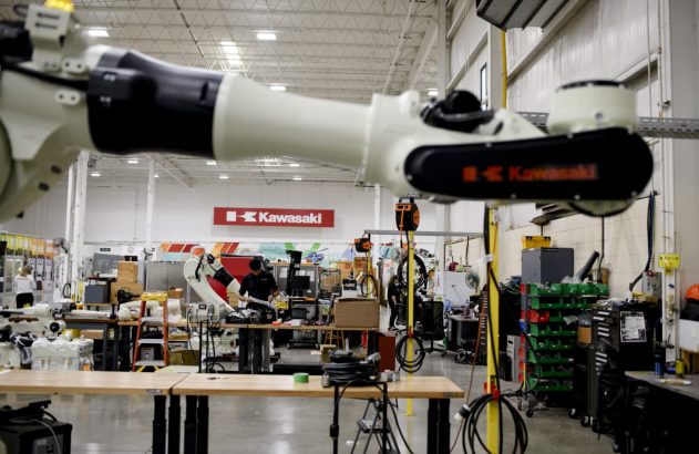 Braço robótico industrial suspenso em fábrica Kawasaki com outros robôs e equipamentos.