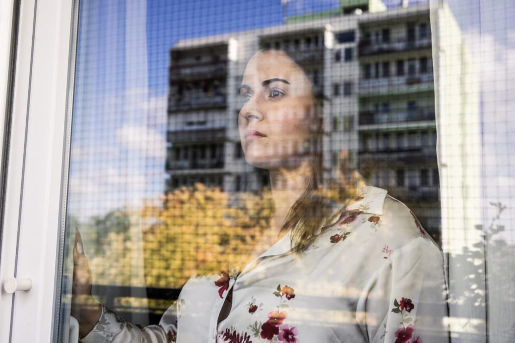 Mulher olha pela janela. No vidro, reflexo de prédio e árvores outonais.