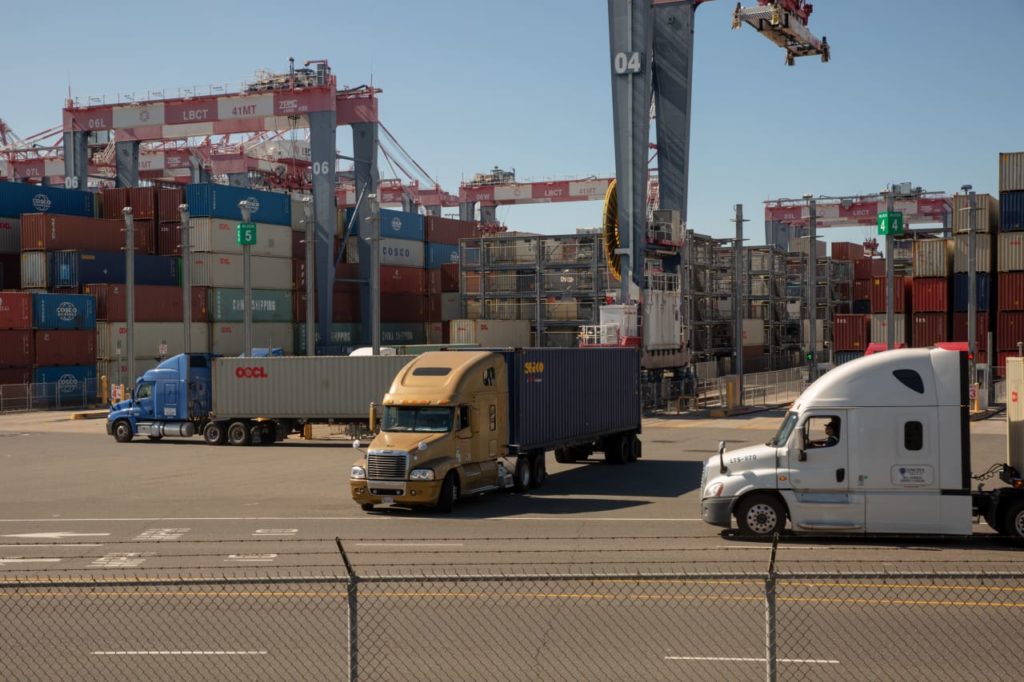 Três carretas em pátio de terminal de contêineres. Pilhas de caixas e guindastes ao fundo. Logística de porto.
