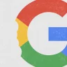 A imagem apresenta o logo estilizado da Google (com um desenho sugerindo uma "mordida"do lado esquerdo), com a letra "G" dividida em segmentos vermelhos, verdes, azuis e amarelos, sobre um fundo cinza claro com pequenas manchas brancas.