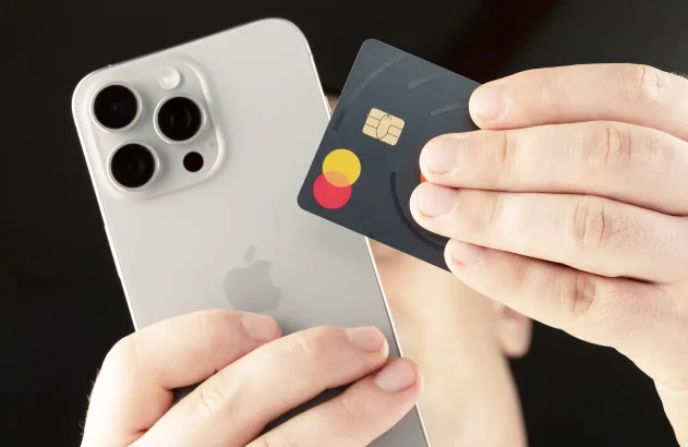 Mãos segurando um iPhone prateado e um cartão Mastercard de design escuro e minimalista.