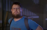 Frame do trailer do documentário “Money Electric: The Bitcoin Mystery”, onde se vê o O desenvolvedor de software Peter Todd, um Homem barbudo com óculos, camiseta azul e mochila preta em ambiente interno.