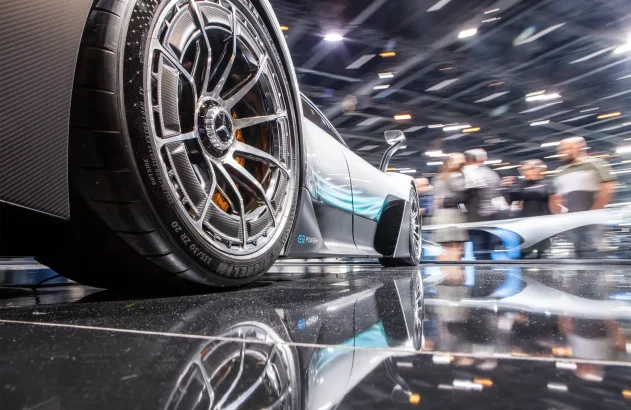 A imagem mostra a roda dianteira esquerda de um carro esportivo de alta performance com aros de liga leve, pneus Michelin e detalhes nas cores prateado, azul e preto, em um salão de automóveis.