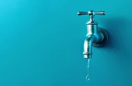 Imagem de uma torneira de metal pingando contra um fundo azul turquesa, com gotas de água prestes a cair.