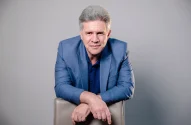 Retrato de Sergio Zimerman, CEO da rede de lojas Petz, de cabelos grisalhos, vestido com blazer e polo azul, apoiado em cadeira bege, em fundo neutro.