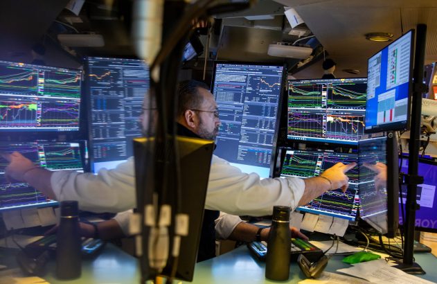 Homem analisando e apontando para múltiplas telas com gráficos financeiros em uma sala de operações.