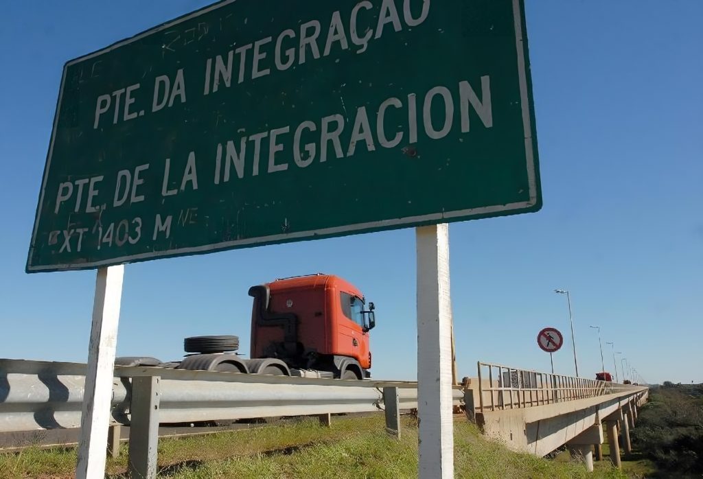 Placa verde indicando 'Ponte da Integração' em português e espanhol; caminhão laranja atravessando a ponte.