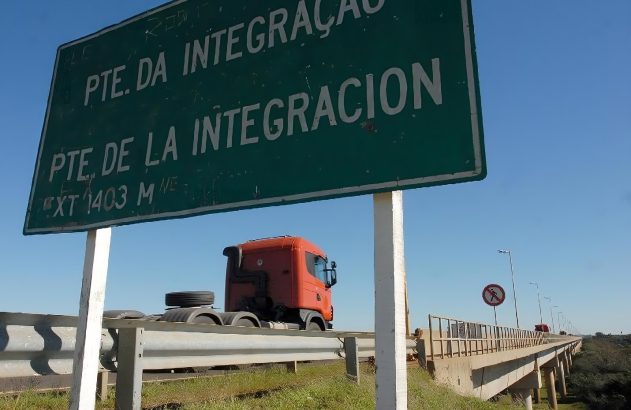 Placa verde indicando 'Ponte da Integração' em português e espanhol; caminhão laranja atravessando a ponte.
