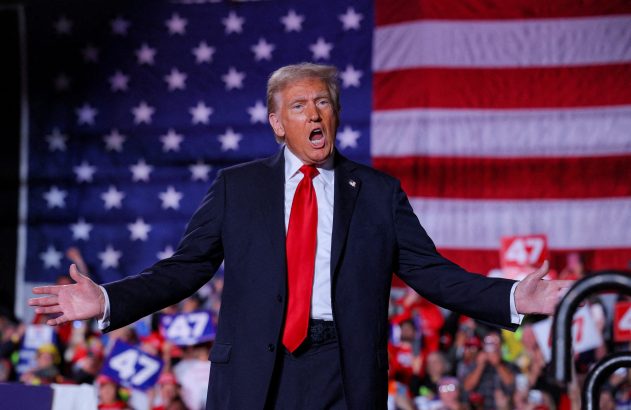 Donald Trump, de terno e gravata, gesticula com braços abertos. Bandeira dos EUA e público.