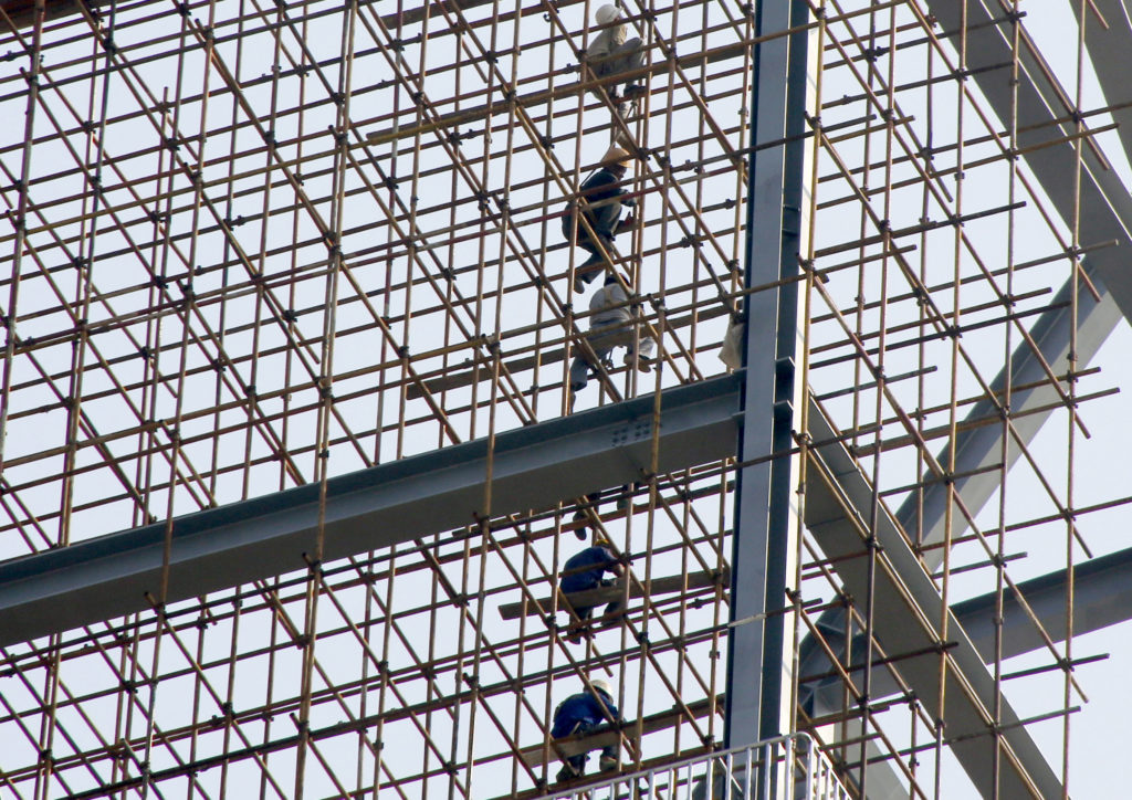 Operários de construção trabalham em andaimes de bambu conectados a uma estrutura metálica.