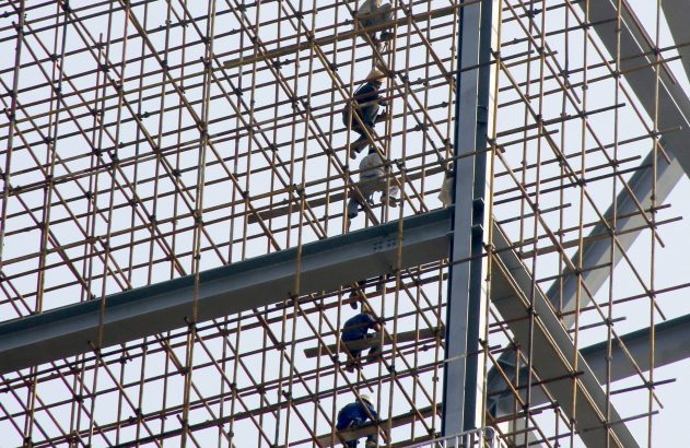 Operários de construção trabalham em andaimes de bambu conectados a uma estrutura metálica.
