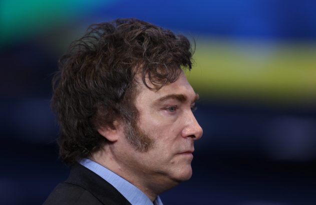 Homem de perfil, cabelo escuro encaracolado e costeletas, olhando à direita. Veste camisa azul e paletó. Fundo colorido borrado.