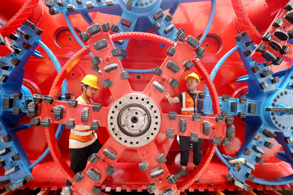 Homens de capacete trabalham dentro de engrenagens gigantes vermelhas e azuis.