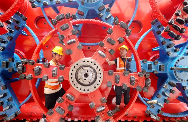 Homens de capacete trabalham dentro de engrenagens gigantes vermelhas e azuis.