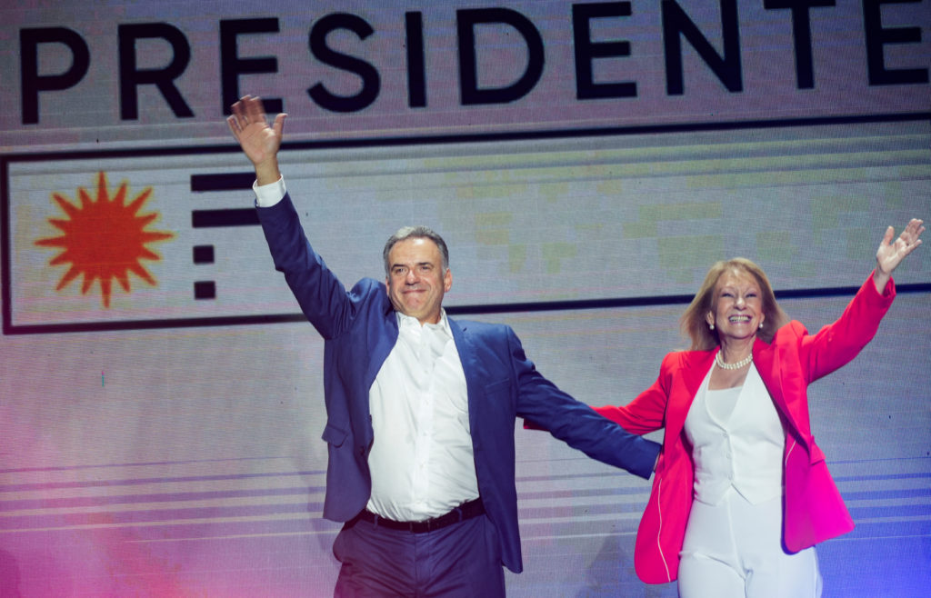 Homem e mulher sorrindo e acenando em palco. Fundo: "PRESIDENTE" e o Sol de Maio uruguaio.