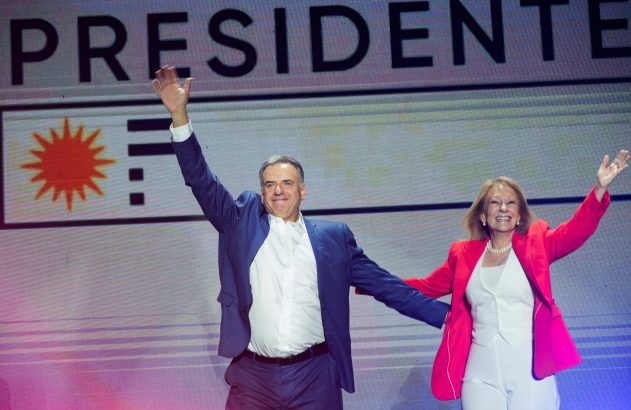 Homem e mulher sorrindo e acenando em palco. Fundo: "PRESIDENTE" e o Sol de Maio uruguaio.