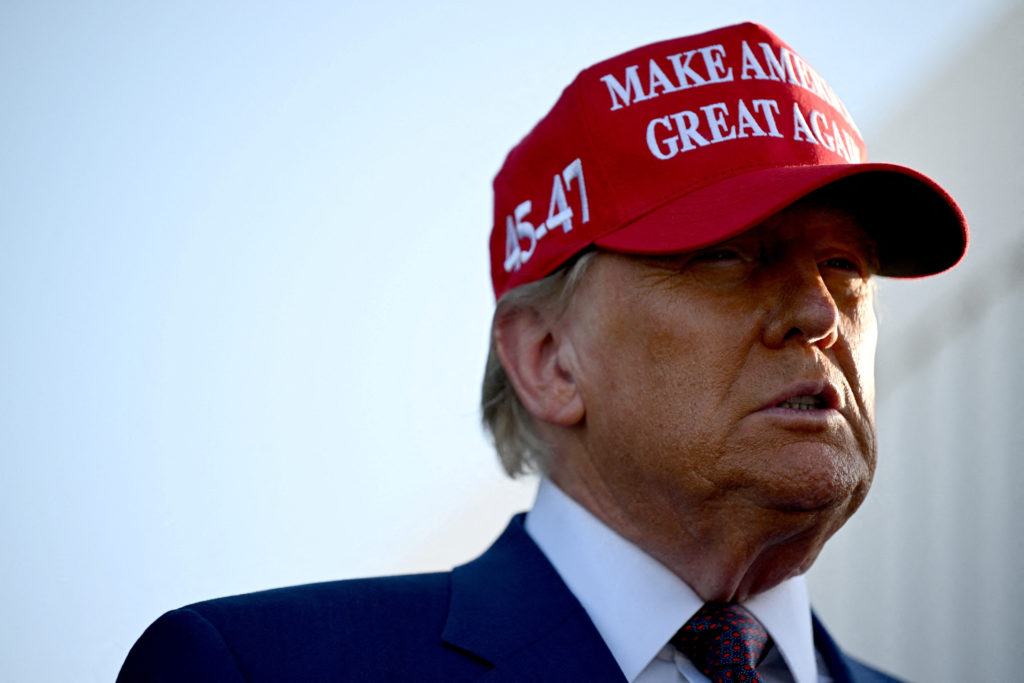 Donald Trump de boné vermelho "Make America Great Again" e "45-47", terno azul, com boca semiaberta.