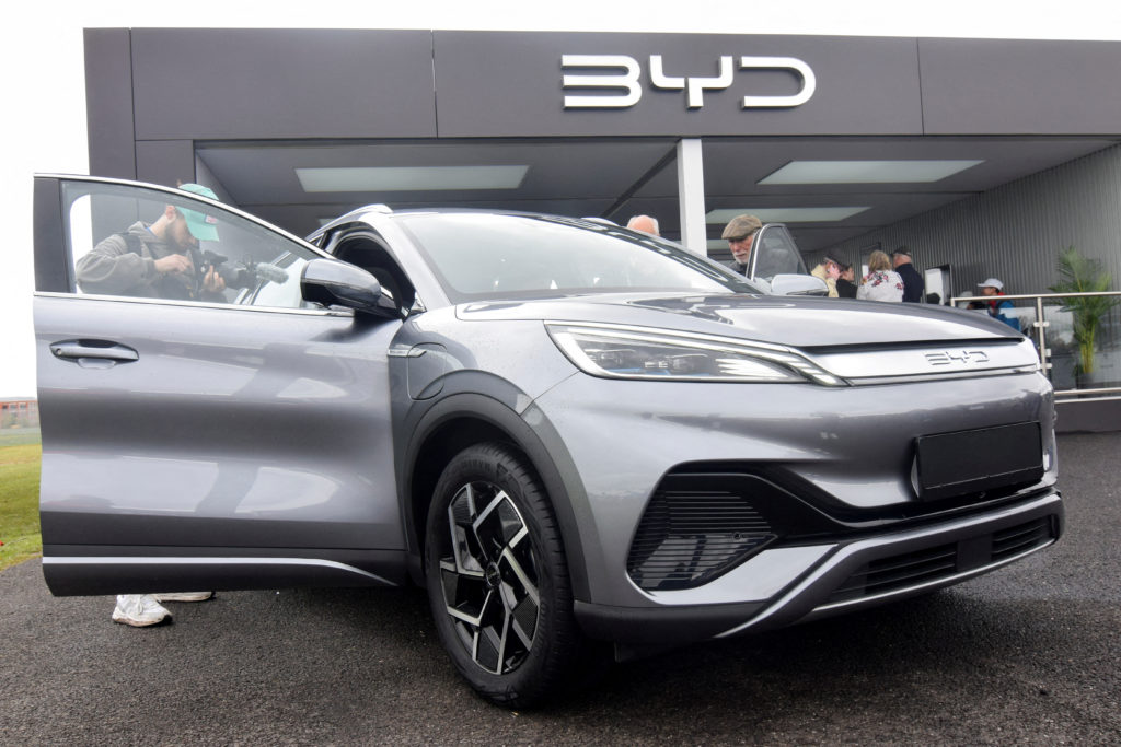 SUV BYD cinza em exibição com porta aberta e pessoa filmando. Prédio com logo BYD e outras pessoas ao fundo.