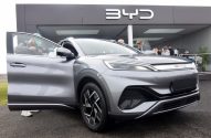 SUV BYD cinza em exibição com porta aberta e pessoa filmando. Prédio com logo BYD e outras pessoas ao fundo.