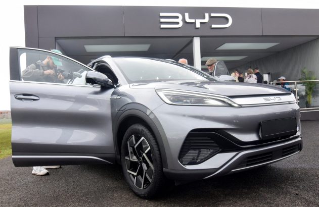 SUV BYD cinza em exibição com porta aberta e pessoa filmando. Prédio com logo BYD e outras pessoas ao fundo.