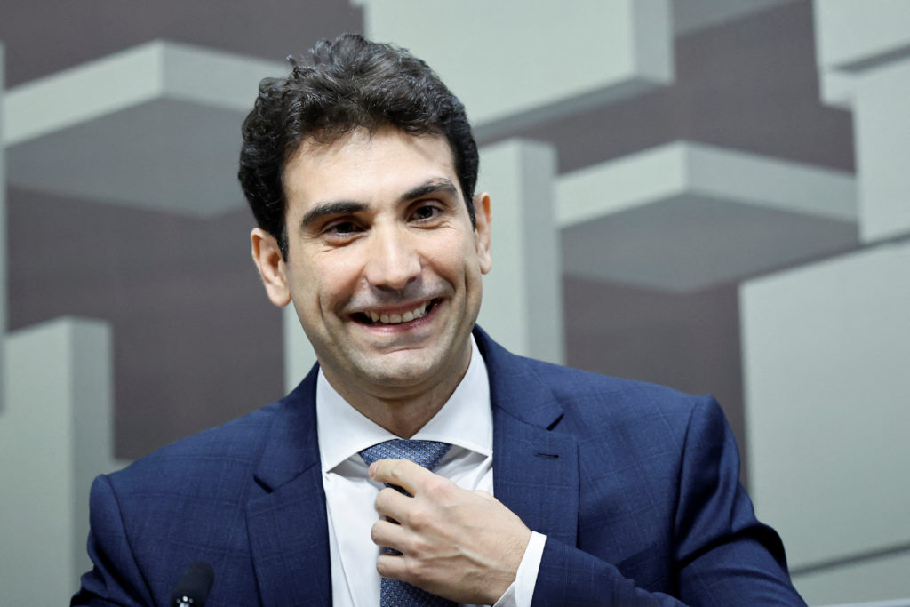 Homem de terno azul sorri e ajusta a gravata. Fundo com formas geométricas cinzas.
