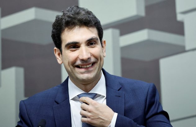 Homem de terno azul sorri e ajusta a gravata. Fundo com formas geométricas cinzas.