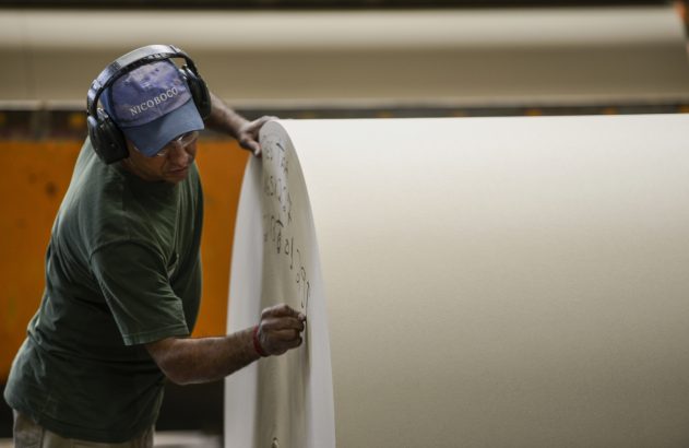 Operário de fábrica, com fones, boné e óculos, escreve em enorme rolo de papel.