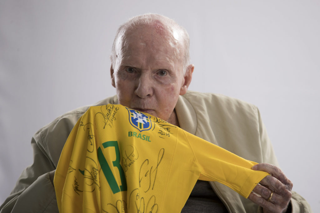 Homem idoso segura camisa autografada da seleção brasileira e olha para a câmera.